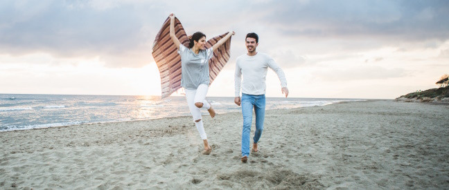 Frau mit Handtuch und Mann laufen lachend den Strand entlang