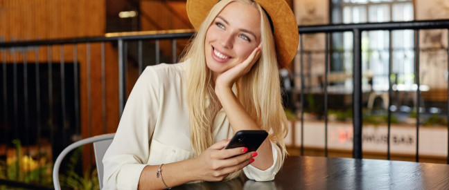 Frau sitzt mit Smartphone im Café und schaut lächelnd in die Ecke