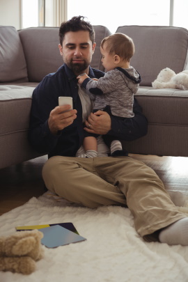 Vater sitzt mit Kind im Wohnzimmer und schaut aufs Smartphone